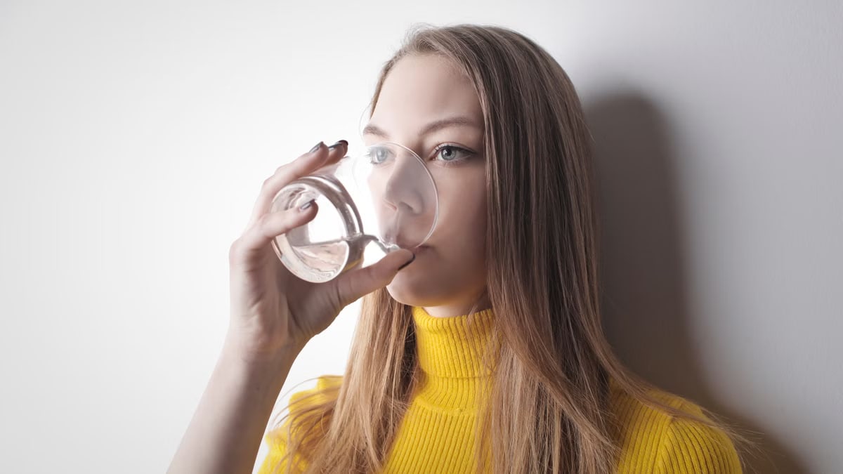 Young woman in a yellow top, drinking water from a glass against a white wall.